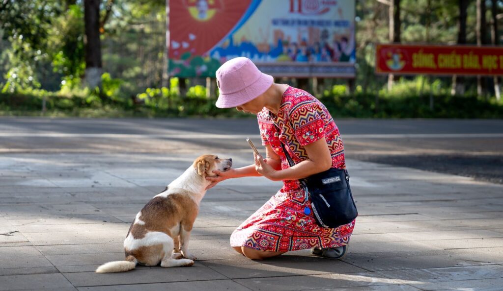 A woman in a colorful dress and bucket hat pets a dog on a sunlit outdoor path.