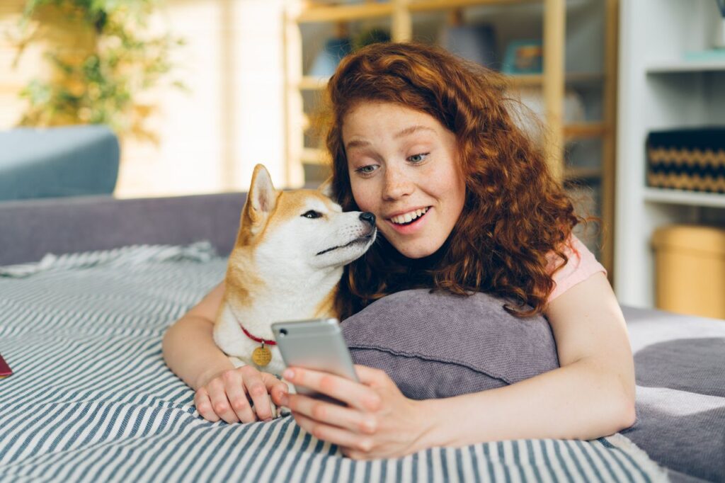 A woman with red hair and her Shiba Inu dog lying on a bed, looking at a smartphone happily.